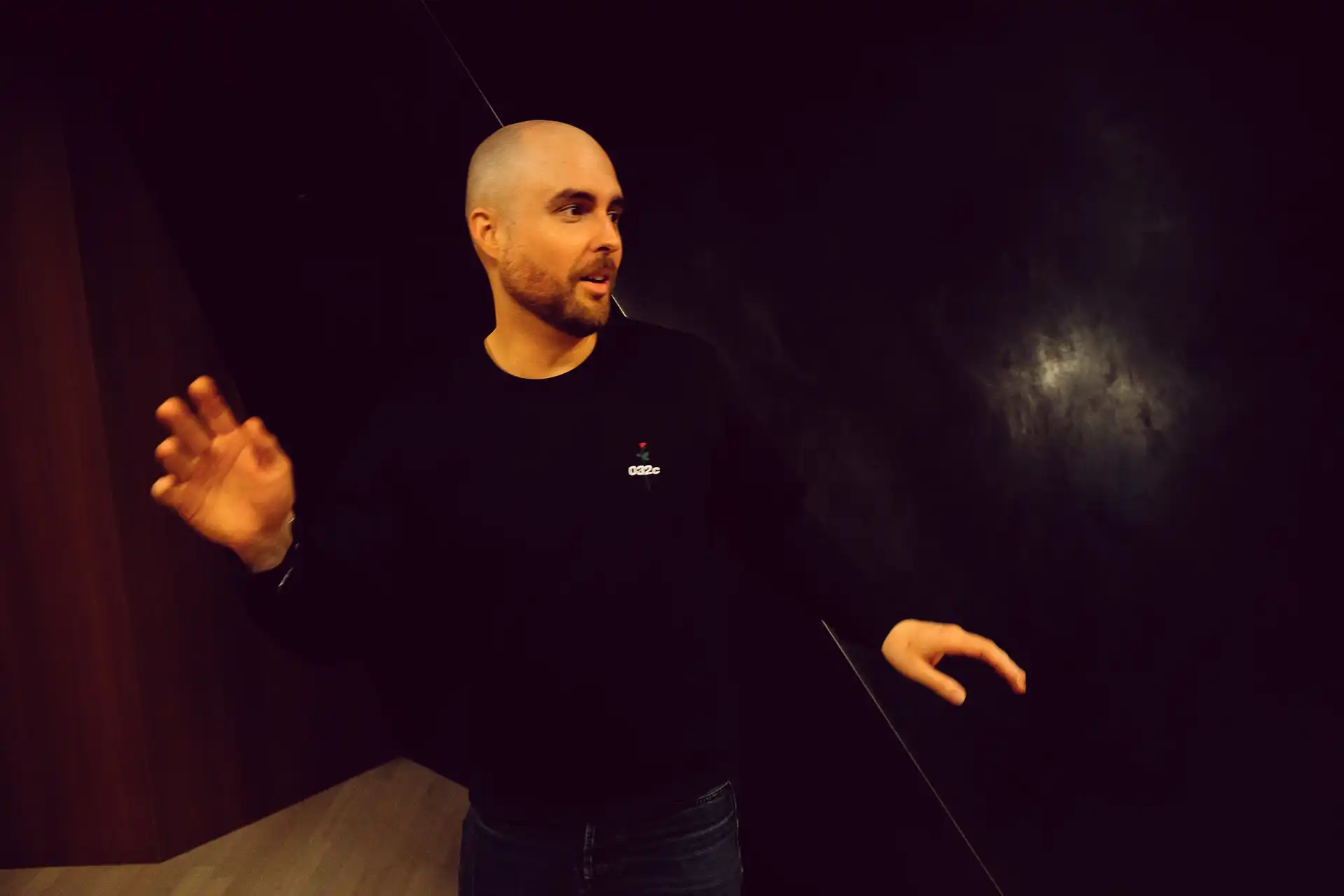 Cyrill Kuhlmann standing in a dimly lit room, gesturing with both hands while looking to the side.